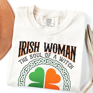 以下が含まれることがあります： 黒とオレンジ色の文字で「IRISH WOMAN THE SOUL OF A WITCH」と書かれたクリーム色のTシャツ。緑とオレンジのシャムロックのデザインは、ケルトノットのボーダーで囲まれています。Tシャツは折りたたまれています。