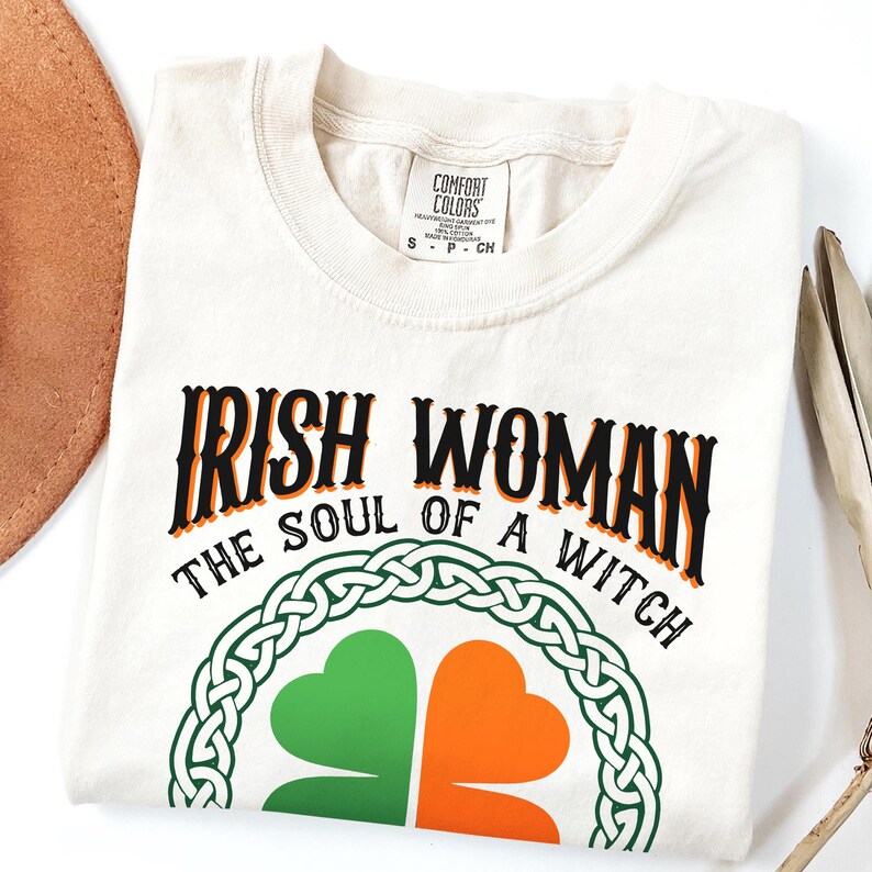 以下が含まれることがあります： 黒とオレンジ色の文字で「IRISH WOMAN THE SOUL OF A WITCH」と書かれたクリーム色のTシャツ。緑とオレンジのシャムロックのデザインは、ケルトノットのボーダーで囲まれています。Tシャツは折りたたまれています。