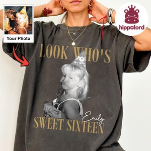 Può includere: Una t-shirt grigio antracite a maniche corte con una fotografia in bianco e nero di un bambino che indossa un cappellino da festa. Il testo "LOOK WHO'S" è sopra la foto e "SWEET SIXTEEN" sotto, con il nome "Emily" scritto in corsivo a destra.