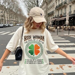 以下が含まれることがあります： 「IRISH WOMAN THE SOUL OF A WITCH」の文字と、緑とオレンジの四つ葉のクローバーのデザインが施されたクリーム色のTシャツ。Tシャツには「THE FIRE OF A LIONESS」、「THE HEART OF A HIPPIE」、「THE MOUTH OF A SAILOR」のフレーズも。ベージュのベースボールキャップが着用されています。