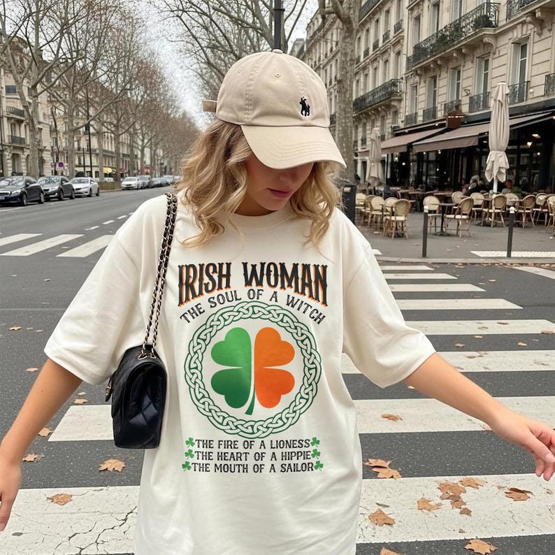 以下が含まれることがあります： 「IRISH WOMAN THE SOUL OF A WITCH」の文字と、緑とオレンジの四つ葉のクローバーのデザインが施されたクリーム色のTシャツ。Tシャツには「THE FIRE OF A LIONESS」、「THE HEART OF A HIPPIE」、「THE MOUTH OF A SAILOR」のフレーズも。ベージュのベースボールキャップが着用されています。