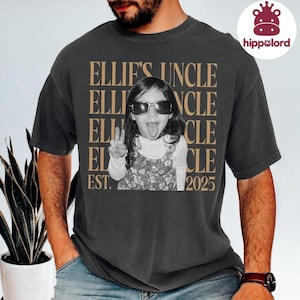 May include: A charcoal gray short-sleeve t-shirt featuring a graphic print of a child wearing sunglasses, making a peace sign, and sticking out their tongue. The text reads ELLIE'S UNCLE and EST. 2025 in a large, tan, repeating serif font.