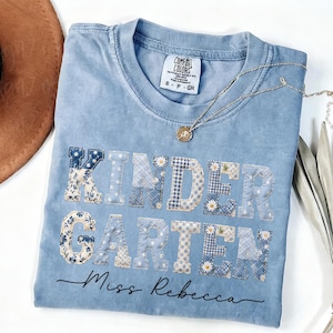 Peut inclure: Un t-shirt bleu clair à col rond avec le mot KINDERGARTEN en grandes lettres style patchwork aux motifs floraux, à carreaux et à pois bleus et blancs. Sous le texte, le nom Miss Rebecca est imprimé en écriture cursive noire.