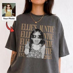 Peut inclure: Un t-shirt gris anthracite &agrave; manches courtes avec une impression graphique personnalis&eacute;e. Le motif comprend le texte r&eacute;p&eacute;t&eacute; "ELLIE'S AUNTIE" en beige, une photo en noir et blanc d'un enfant portant des lunettes de soleil et faisant un signe de paix.