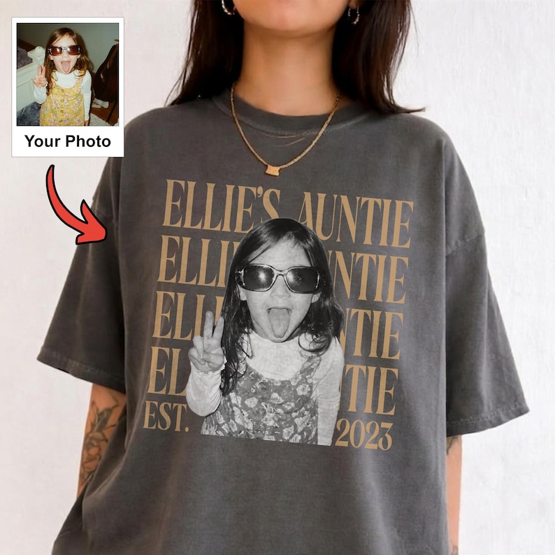 Peut inclure: Un t-shirt gris anthracite &agrave; manches courtes avec une impression graphique personnalis&eacute;e. Le motif comprend le texte r&eacute;p&eacute;t&eacute; "ELLIE'S AUNTIE" en beige, une photo en noir et blanc d'un enfant portant des lunettes de soleil et faisant un signe de paix.