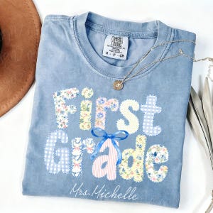 May include: A blue short-sleeved t-shirt with a crew neckline featuring the text First Grade in multi-colored patterned letters, including floral, gingham, and striped designs. A blue ribbon graphic sits above the letter a, and Mrs. Michelle is printed below.