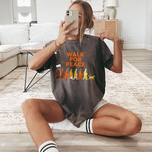 Op de afbeelding: Donkergrijs t-shirt met de oranje tekst "WALK FOR PEACE". Daaronder staat een afbeelding van mensen die in een rij lopen, met een hond aan het einde. Het t-shirt wordt door een persoon gedragen.