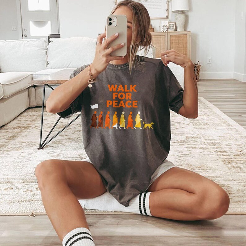 Op de afbeelding: Donkergrijs t-shirt met de oranje tekst "WALK FOR PEACE". Daaronder staat een afbeelding van mensen die in een rij lopen, met een hond aan het einde. Het t-shirt wordt door een persoon gedragen.