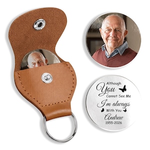 May include: A tan leather keychain pouch with a silver snap button and a metal ring. Inside the pouch is a circular metal insert featuring a photograph of an older man. A separate circular metal disc displays the text: Although You Cannot See Me I'm always With You Andrew 1955-2026, accompanied by two small butterfly icons.