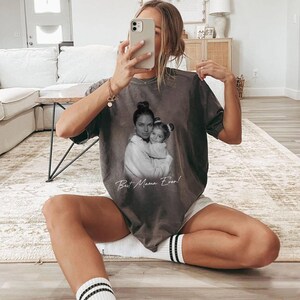 Pu&ograve; includere: Una maglietta grigia con un'immagine in bianco e nero di una madre e un bambino che si abbracciano. Il testo "Best Mama Ever!" &egrave; stampato sotto. La maglietta &egrave; indossata.
