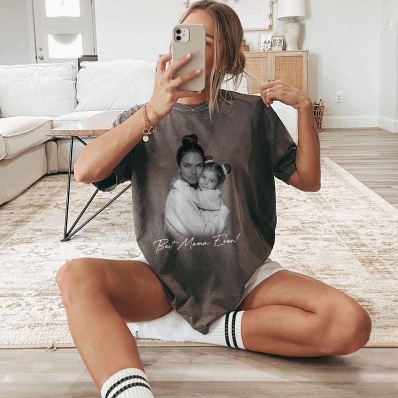 Pu&ograve; includere: Una maglietta grigia con un'immagine in bianco e nero di una madre e un bambino che si abbracciano. Il testo "Best Mama Ever!" &egrave; stampato sotto. La maglietta &egrave; indossata.