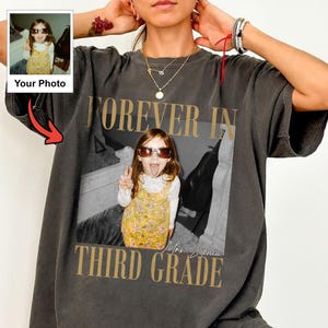 Może przedstawiać: Ciemnoszary t-shirt z krótkim rękawem z dużym prostokątnym nadrukiem przedstawiającym dziecko w okularach przeciwsłonecznych i żółtej sukience w kwiaty. Napis FOREVER IN THIRD GRADE jest nadrukowany złotą czcionką nad i pod zdjęciem.