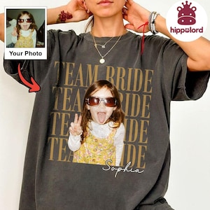 May include: A charcoal gray short-sleeve t-shirt featuring a large, repeating gold-toned text graphic that reads TEAM BRIDE. Centered on the shirt is a photographic print of a child wearing brown sunglasses and a yellow floral dress.