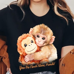 Könnte beinhalten: Schwarzes T-Shirt mit einer Aquarellillustration von zwei sich umarmenden Affen. Ein Affe ist hellbraun, der andere orange. Der Text "You Are Never Alone, Punch!" ist in kursiver Schrift unter dem Bild gedruckt.