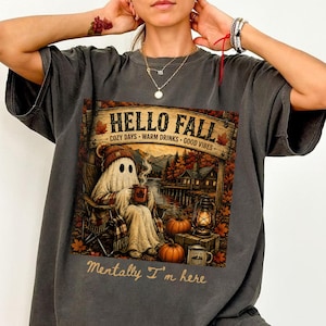 May include: A charcoal gray t-shirt featuring a graphic of a ghost in a beanie sitting in a chair by a lake. The text reads HELLO FALL COZY DAYS WARM DRINKS GOOD VIBES and Mentally I'm here. The scene includes pumpkins, a lantern, and a cabin.