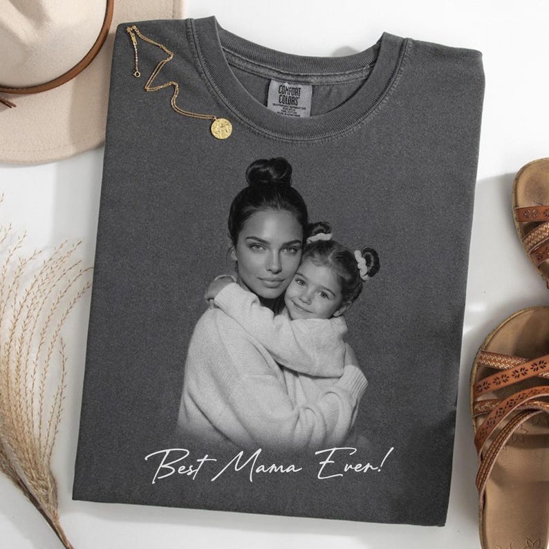 Pu&ograve; includere: T-shirt grigio scuro con un'immagine in bianco e nero di una madre e un bambino che si abbracciano. Il testo "Best Mama Ever!" &egrave; stampato in bianco. Una collana d'oro &egrave; appoggiata sulla maglietta.