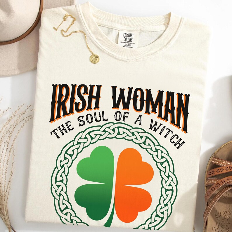 以下が含まれることがあります： 黒とオレンジ色の「IRISH WOMAN THE SOUL OF A WITCH」という文字が入ったクリーム色のTシャツ。緑とオレンジの四つ葉のクローバーのデザインがテキストの下に配置され、緑のケルトノットのボーダーで囲まれています。金のネックレスがシャツの上にかけられています。