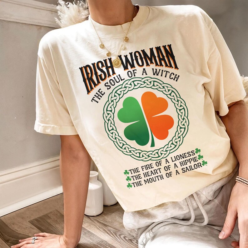以下が含まれることがあります： 「IRISH WOMAN THE SOUL OF A WITCH」の文字と、緑とオレンジのクローバーのデザインが施されたクリーム色のTシャツ。「THE FIRE OF A LIONESS THE HEART OF A HIPPIE THE MOUTH OF A SAILOR」の文字も含まれています。