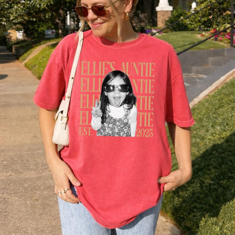 Peut inclure: Un t-shirt rouge &agrave; manches courtes avec une photographie en noir et blanc d'un enfant portant des lunettes de soleil et tirant la langue. Le texte "ELLIE'S AUNTIE" est r&eacute;p&eacute;t&eacute; en arri&egrave;re-plan en dor&eacute;, avec "EST. 2023" imprim&eacute; en bas.