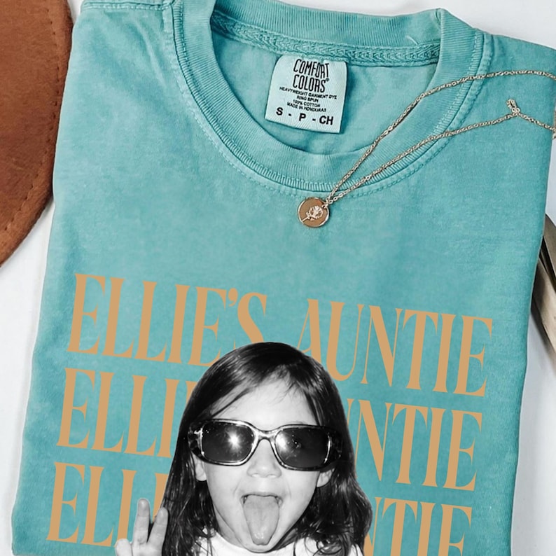 Peut inclure: Un t-shirt &agrave; manches courtes bleu sarcelle avec un col rond et une &eacute;tiquette Comfort Colors. Le t-shirt pr&eacute;sente le texte orange "ELLIE'S AUNTIE" r&eacute;p&eacute;t&eacute; trois fois et une photographie en noir et blanc d'un enfant portant des lunettes de soleil.