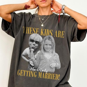 Op de afbeelding: Een antracietgrijs t-shirt met korte mouwen en een zwart-witfoto van twee kinderen. De tekst "THESE KIDS ARE" staat boven de foto en "Alex & Emily GETTING MARRIED!" is in gouden letters onder de afbeelding gedrukt.