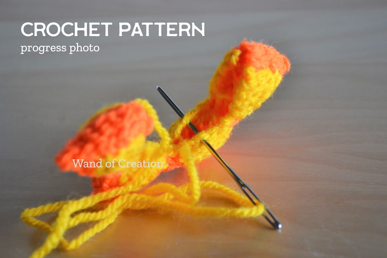 May include: A close-up of a crochet project in progress. The project uses yellow and orange yarn, with two needles inserted. The text "CROCHET PATTERN progress photo" and "Wand of Creation" are visible.
