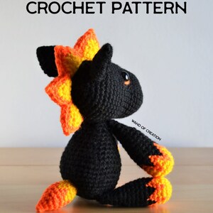 May include: A black crochet creature with orange and yellow detailing, including a spiky mane, paws, and tail. The text "CROCHET PATTERN" is displayed above the creature. The creature is sitting on a wooden surface.