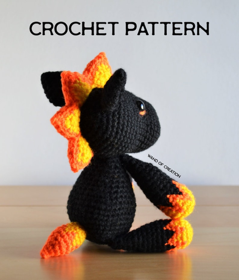 May include: A black crochet creature with orange and yellow detailing, including a spiky mane, paws, and tail. The text "CROCHET PATTERN" is displayed above the creature. The creature is sitting on a wooden surface.