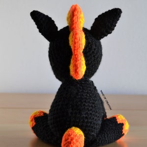 May include: A crocheted unicorn toy in black, featuring orange and yellow details. The mane and tail are in vibrant orange and yellow. The image includes the text "CROCHET PATTERN" at the top.