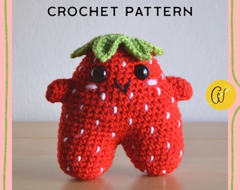 Puede incluir: Un juguete de peluche de fresa tejido a crochet con una parte superior verde de hojas, ojos negros y una boca sonriente. El juguete es rojo con detalles blancos y tiene una expresión amigable.