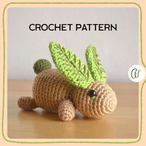 May include: Crochet pattern for a brown bunny with green leaf ears. The bunny is lying on a wooden surface. The pattern is called "Crochet Pattern".