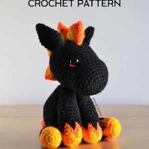 May include: A black crochet dragon with orange and yellow details. The dragon has large black eyes and is sitting on a wooden surface. The text "CROCHET PATTERN" is at the top of the image.