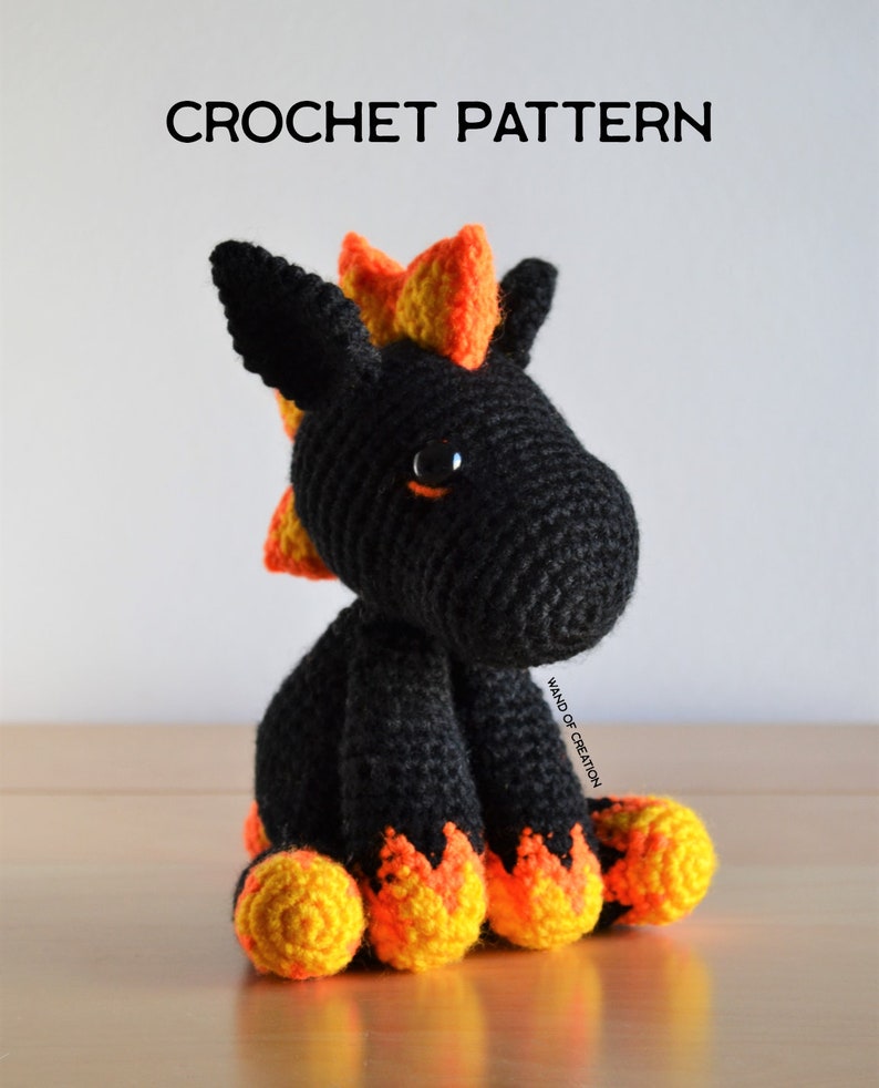 May include: A black crochet dragon with orange and yellow details. The dragon has large black eyes and is sitting on a wooden surface. The text "CROCHET PATTERN" is at the top of the image.