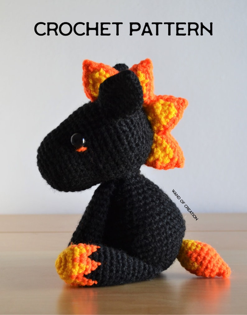 May include: A crocheted dragon toy, predominantly black with orange and yellow detailing. The dragon features a spiky mane and tail. The image includes the text "CROCHET PATTERN" at the top.