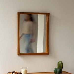 Puede incluir: Una mesa consola de madera con un estante y un espejo colgado encima. La mesa tiene un acabado de madera clara y el espejo tiene un marco rectangular simple.