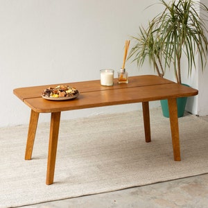 以下が含まれることがあります： 明るい茶色の木製のコーヒーテーブルで、天板が分割され、両側に3本の脚があります。テーブルは薄いベージュの敷物の上に置かれています。テーブルの上には、スナックの入った皿と、キャンドルとディフューザーがあります。