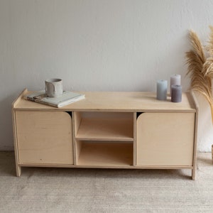 Puede incluir: Un armario de madera clara con dos puertas y dos estantes. El armario tiene bordes redondeados y está sobre una alfombra beige claro. Hay tres velas y una taza de cerámica en la parte superior del armario.