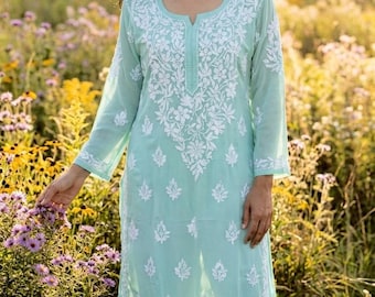 Puede incluir: Un vestido túnica turquesa claro con intrincados bordados florales blancos. El vestido de manga larga presenta un cuello redondo y una abertura en el dobladillo. La mujer está de pie en un campo de flores silvestres.
