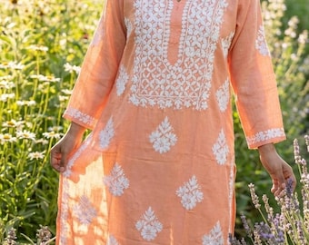 Puede incluir: Túnica color melocotón con bordados florales blancos. La túnica de manga larga presenta un cuello redondo con un pequeño escote en V y se combina con pantalones color crema. El bordado se concentra alrededor del cuello y se dispersa por la túnica.