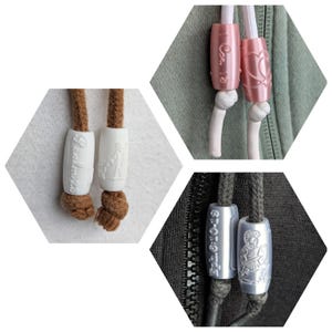 Puede incluir: Primer plano de tres extremos de cordones de sudaderas con capucha diferentes. Uno es blanco con una figura religiosa, otro es rosa con un corazón y el último es plateado con un soldado romano. Cada uno tiene un diseño único.