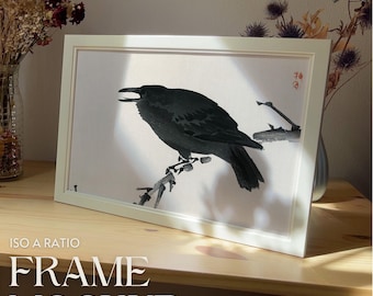 May include: A white frame with a black crow perched on a branch. The crow is facing the right side of the frame. The background is a light beige color. The frame is sitting on a wooden surface.