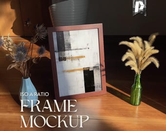 May include: A brown wooden frame with a black and white abstract painting inside. The frame is sitting on a wooden surface with a vase of dried flowers and a bottle of dried grass.