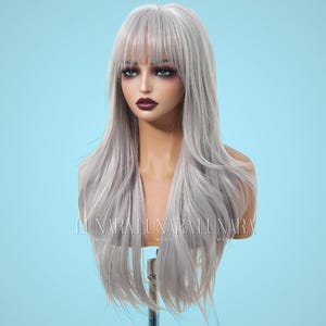 May include: A long, silver-gray wig with a blunt fringe, styled on a mannequin head. The wig has long, flowing layers and is displayed against a light blue backdrop. The text "LUNARA WIGS" is visible on the mannequin.