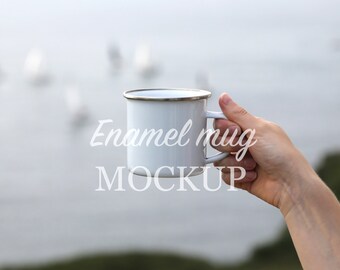 May include: White enamel mug with a silver rim and handle. The mug is held up against a blurred background of water and boats. The text "Enamel mug MOCKUP" is printed on the mug.