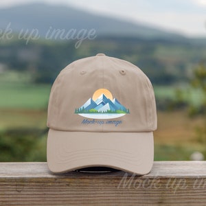 Könnte beinhalten: Beige Baseball Cap mit einem Berg- und Sonnendesign. Die Grafik zeigt blaue Berge, eine gelbe Sonne und grüne Bäume. Der Text "Mock up image" ist auf der Kappe aufgedruckt.