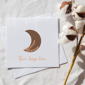 Peut inclure: Une carte blanche avec une lune croissante brune avec des branches dorées et le texte "Your design here". La carte est sur un fond de tissu blanc avec une branche de boules de coton.
