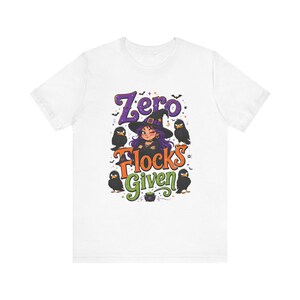 May include: A white short-sleeve t-shirt featuring a graphic print of a winking witch with purple hair wearing a black hat, surrounded by four black birds and a small green cauldron. The text reads Zero FLOCKS Given in purple, orange, and green.