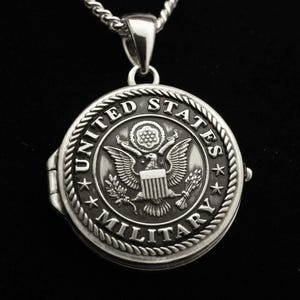 May include: Silver locket necklace with a detailed engraving of the United States Military seal. The seal features an eagle with a shield and olive branches.