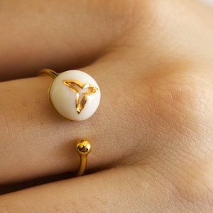 Peut inclure: Bague ouverte dorée avec une pièce maîtresse circulaire blanche. La pièce maîtresse présente un motif floral doré stylisé. La conception de la bague comprend une petite boule dorée à l'extrémité ouverte.