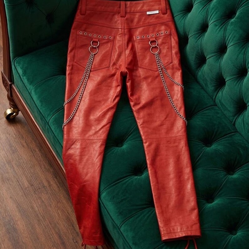 May include: Red leather trousers with silver chain detailing and ring accents on the back pockets. The trousers have a classic five-pocket design and a fitted silhouette. The leather appears to be a matte finish.